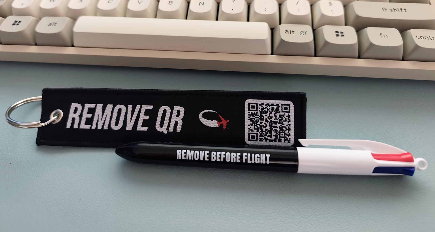 Porte-clés tissé Remove QR scanné sur un bureau, à côté d'un stylo Remove Before Flight
